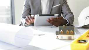 Homem sentado em frete a uma mesa com projetos e EPIs, mexendo no seu tablet e verificando o programa de qualidade na construção