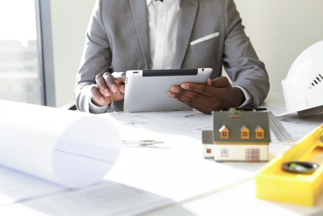 Homem sentado em frete a uma mesa com projetos e EPIs, mexendo no seu tablet e verificando o programa de qualidade na construção