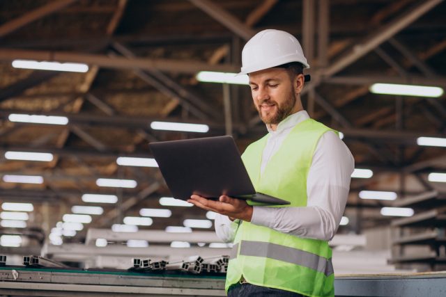 Foto de um homem com colete verde e capacete dentro de uma galpão, segurando um notebook aberto