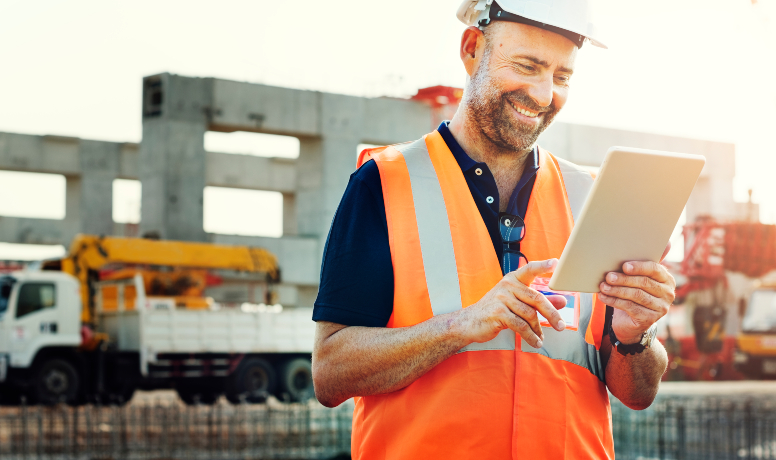 A imagem contém um engenheiro olhando satisfatoriamente para o seu tablet. Ao fundo temos um caminhão estacionado em um canteiro de obras.