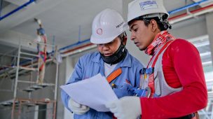 Imagem de dois homens com roupas de trabalho de construção civil, olhando em um papel.