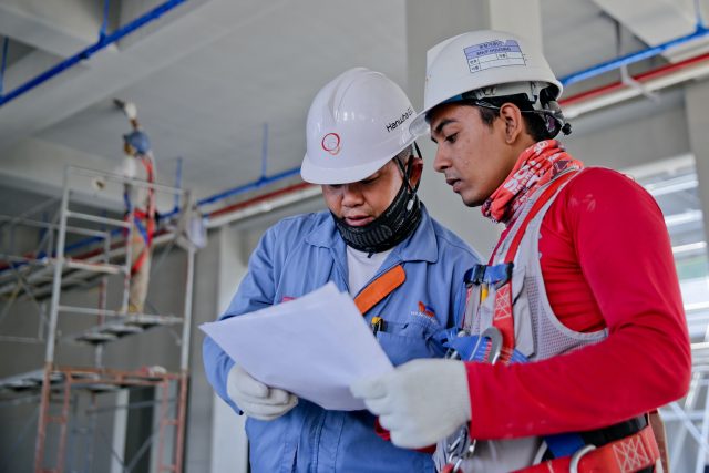 Imagem de dois homens com roupas de trabalho de construção civil, olhando em um papel.