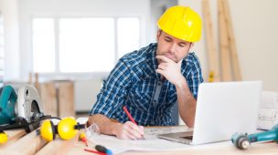 Imagem de um homem com camisa xadrez e capacete amarelo, sentando em frente a uma mesa com uma computador.