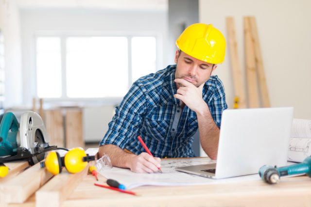 Imagem de um homem com camisa xadrez e capacete amarelo, sentando em frente a uma mesa com uma computador.