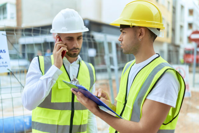 Dois homens com EPIs analisando documentos na obra e falando no celular